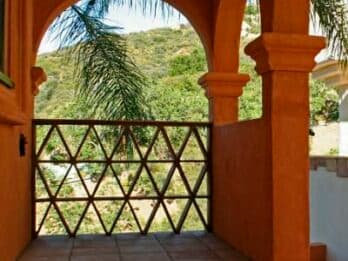 A view from an arched balcony featuring a decorative wooden railing and lush greenery beyond.