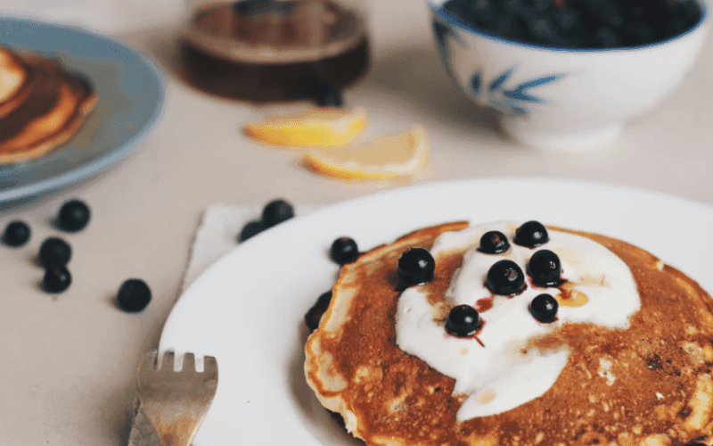 A plate of pancakes topped with yogurt and blueberries, accompanied by lemon slices and a bowl of blueberries in the background. A plate of pancakes topped with yogurt and blueberries, accompanied by lemon slices and a bowl of blueberries in the background.
