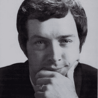 A man with short, wavy hair rests his chin on his hand, smiling softly.