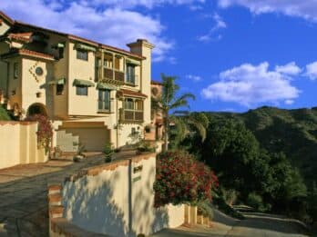 A two-story Mediterranean-style house surrounded by greenery against a blue sky.
